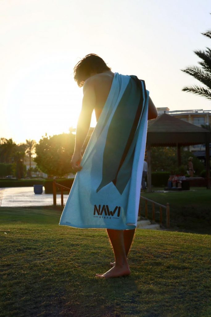 Imagen chico en verano con una toalla Nawi azul con una tabla de surf a la espalda y a contraluz.